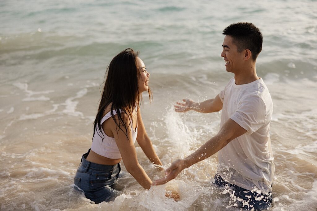A man and a woman kneel in the water of the ocean while on a beach of Oahu and smile at one another while taking engagement photos 