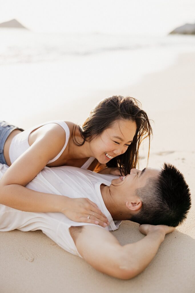 A man lays on a beach and smiles while a woman lays on top of him and smiles back at him as the sun sets behind them