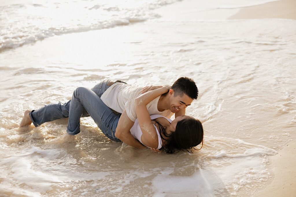 A man and woman wearing jeans and white shirts lay together in the water on a beach of Oahu at sunset while taking their engagement photos