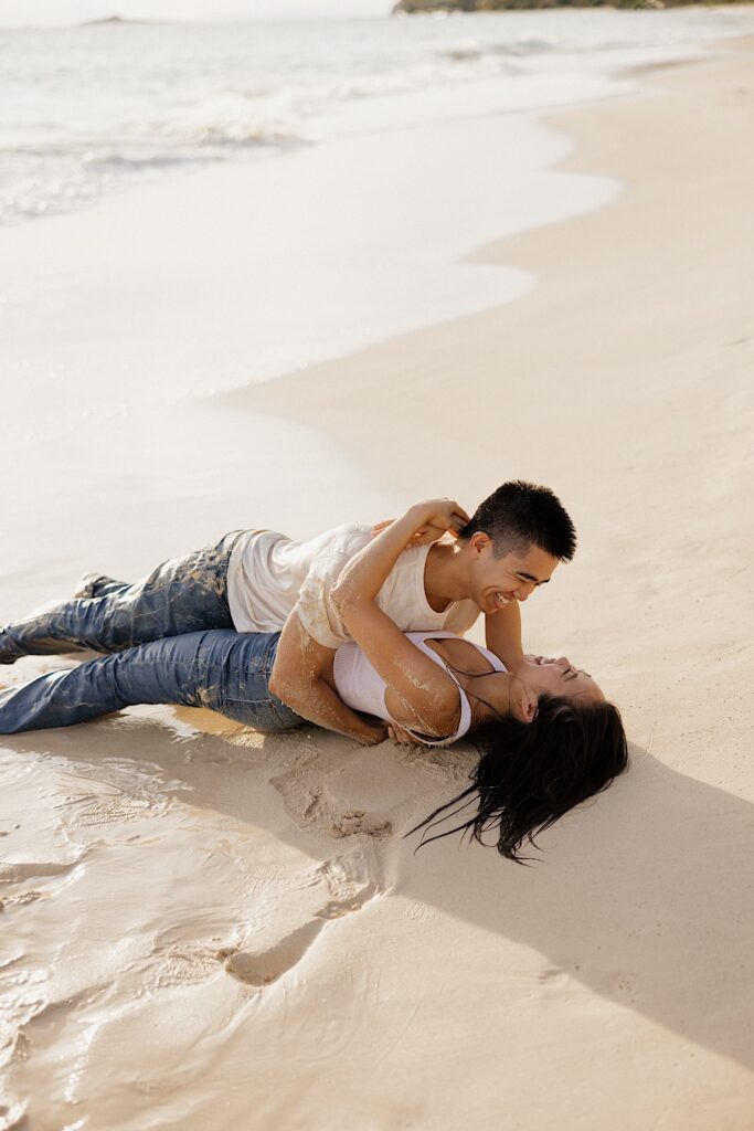 A man and woman lay together on a beach of Oahu and smile as the sun sets behind them and their clothes are wet