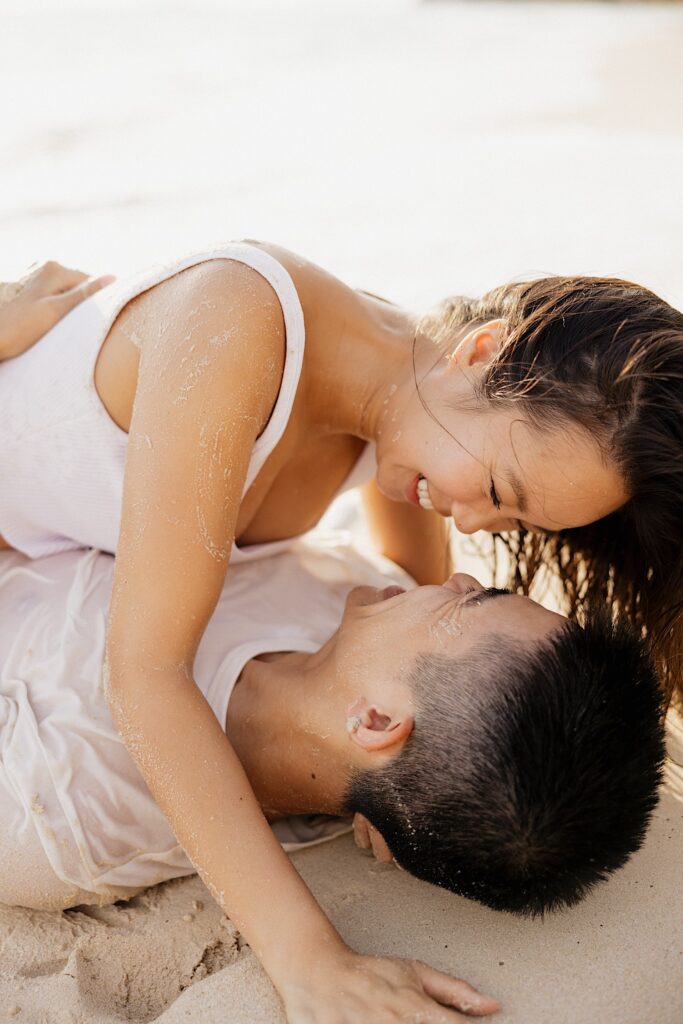 A man and woman wet and covered in sand lay on top of one another and smile