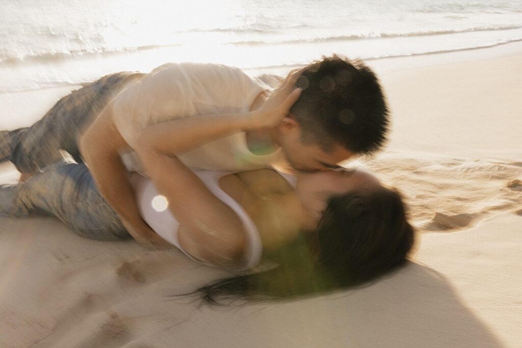 A couple kiss one another while laying on a beach of Oahu as the sun sets behind them while taking their engagement photos