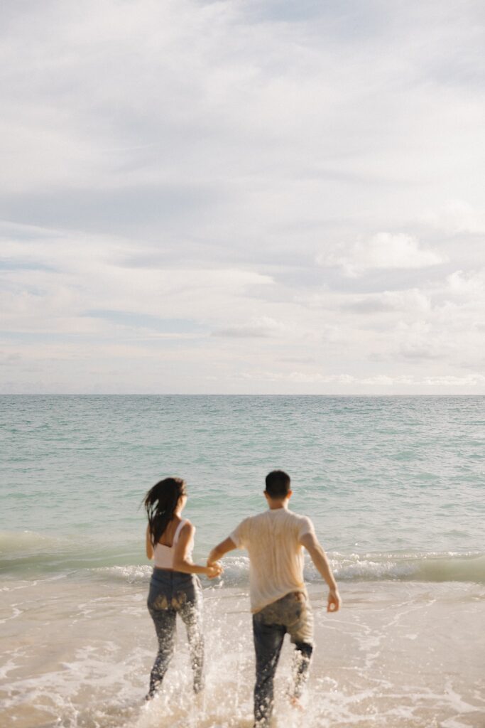 A couple hold hands as they run into the water of the ocean together at sunset