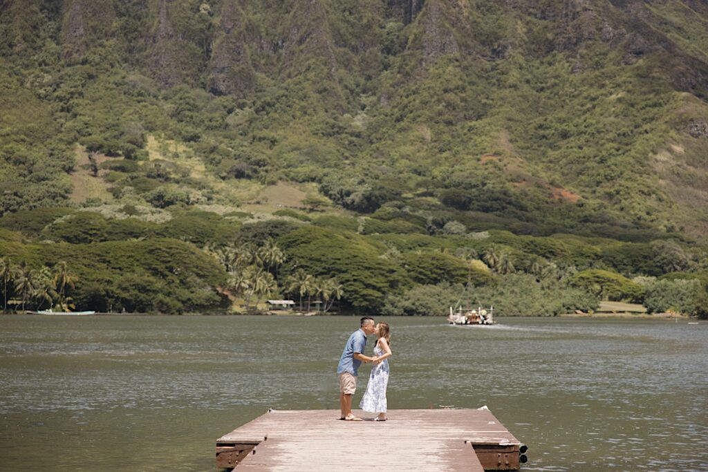 A man and woman kiss while on a dock looking out over a lake in front of a mountain before their surprise proposal on Oahu
