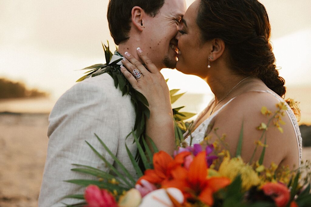 A couple in their wedding attire smile as they're about to kiss while on a beach of Oahu as the sun sets behind them