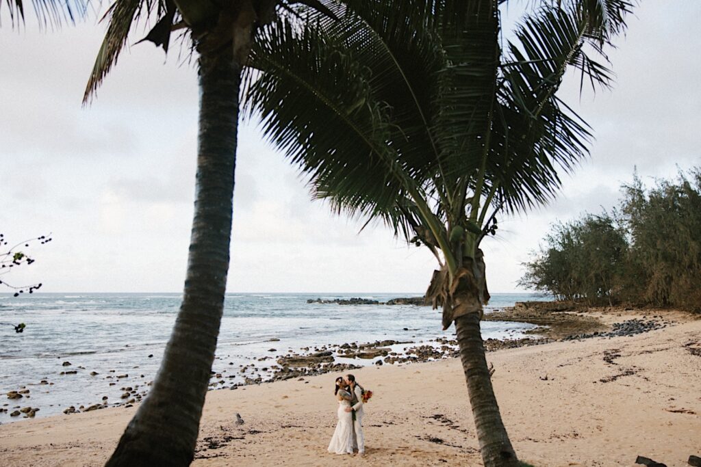A bride and groom kiss while standing on a beach of Oahu with palm trees above them