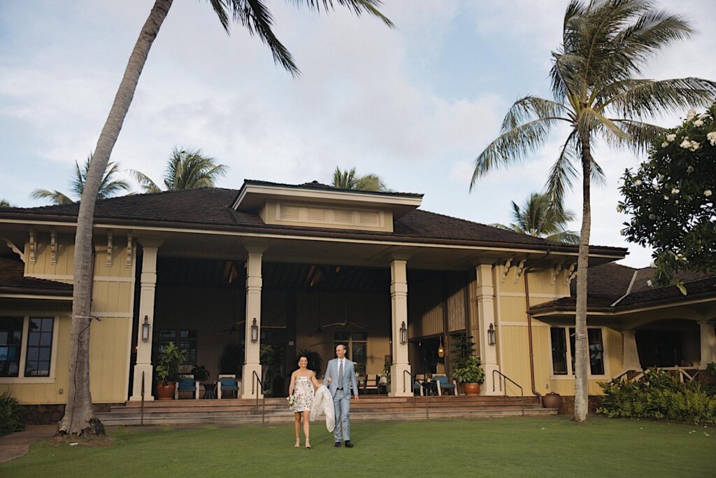 A bride and groom walk hand in hand in front of the clubhouse at Kukui'ula, their wedding venue, towards the outdoor tent for their reception