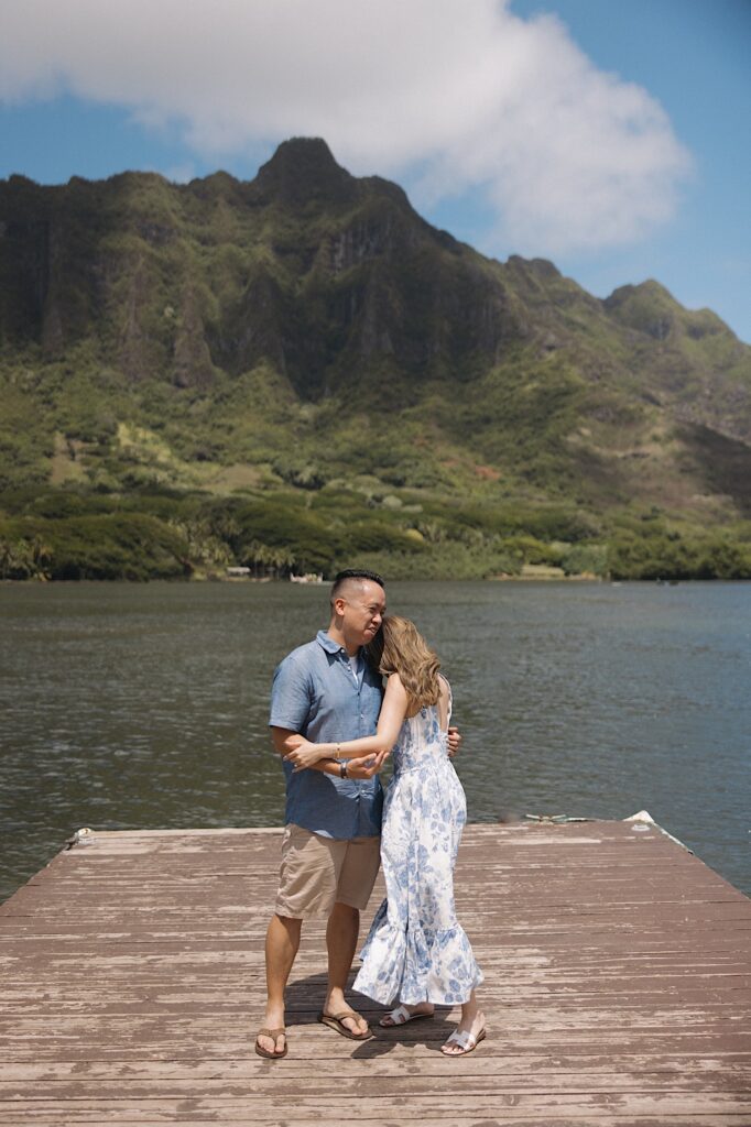 A man and woman embrace one another while standing on a dock in front of a lake and a mountain on Oahu