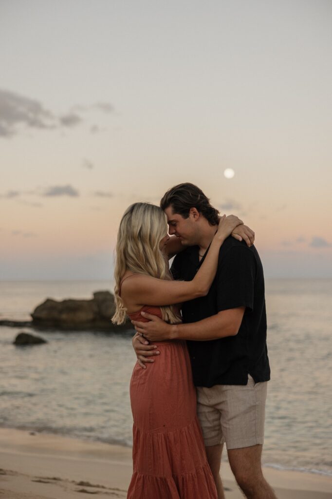 A man and woman embrace and touch foreheads together while on a beach at sunrise as the moon still sits in the sky above them