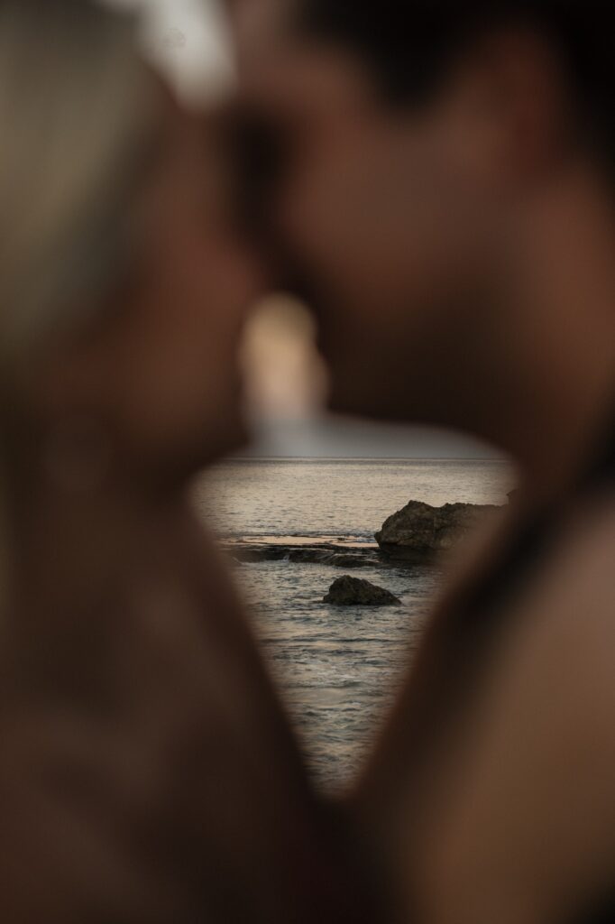 A man and woman smile as they're about to kiss in the foreground of the photo out of focus, in focus behind them are rocks in the ocean