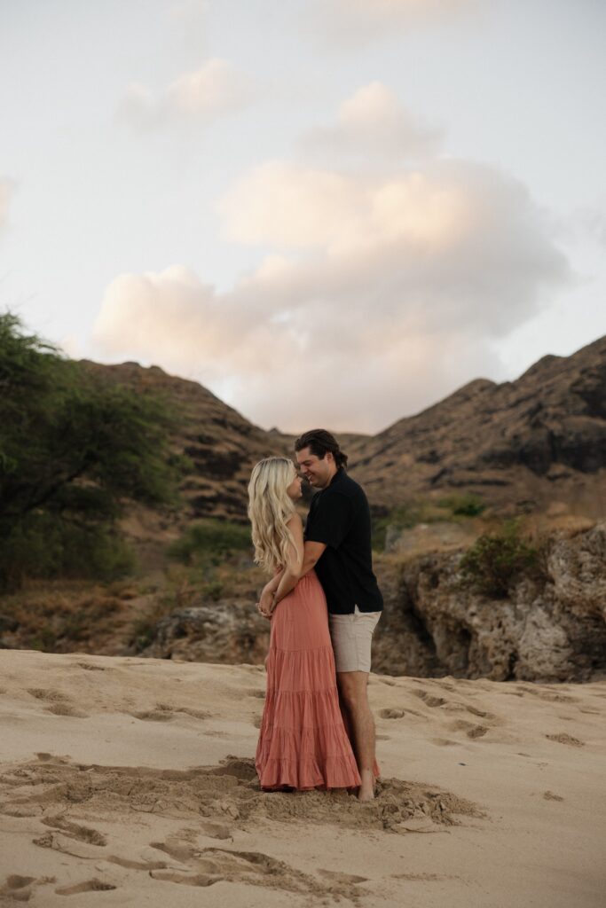 A man and woman hug one another and smile while standing on a beach  with a rocky hill behind them