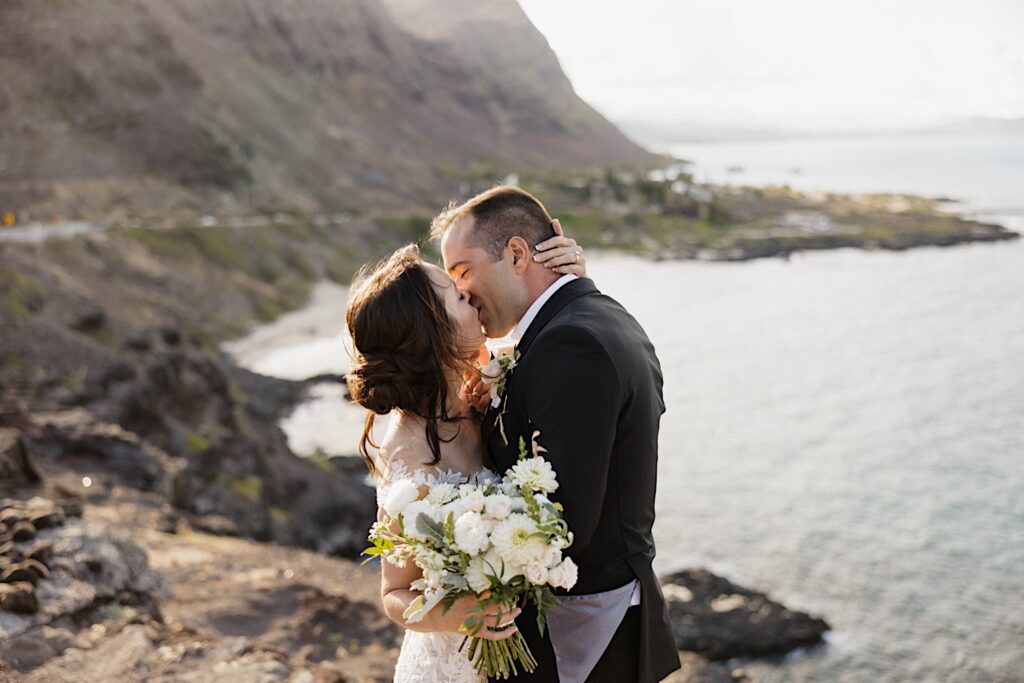 A bride and groom kiss one another while standing atop a cliff on the island of Oahu with the ocean behind them as the sun begins to set