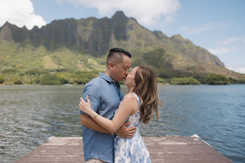 After their surprise proposal a man and woman kiss while standing on a dock in front of a lake and mountains on Oahu