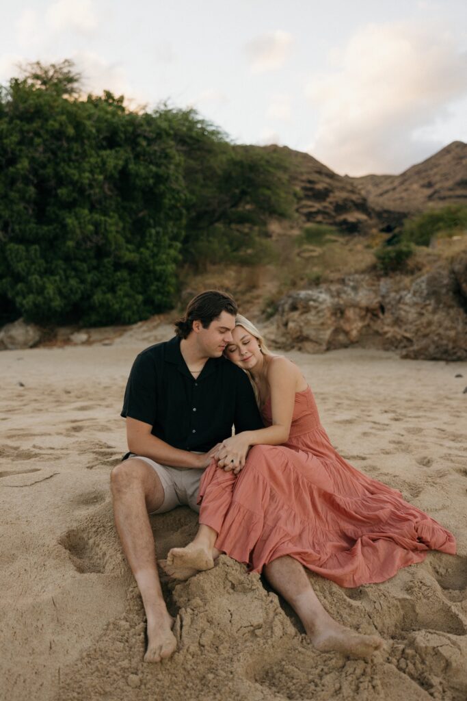 A man and woman sit together on a beach with a rocky hill behind them and snuggle with one another while holding hands