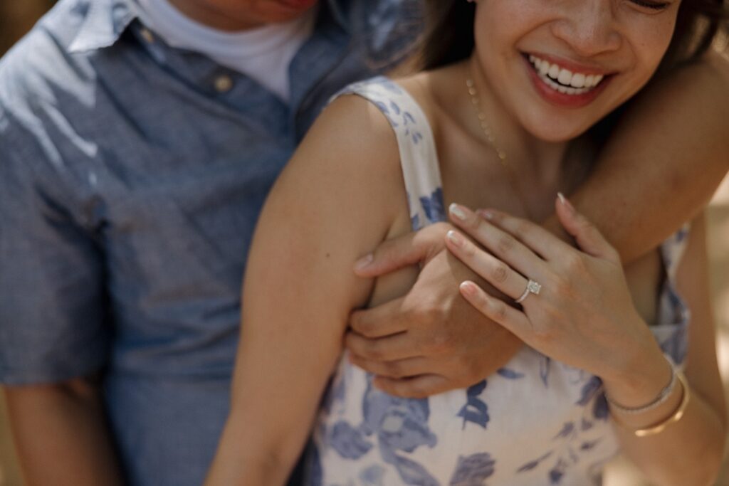 A woman smiles as she's hugged from behind by a man while she shows off her engagement ring after their surprise proposal on Oahu