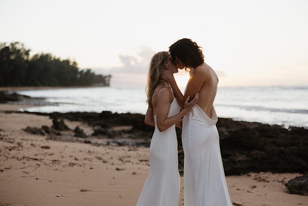 2 brides stand on the beach together and kiss one another as the sun sets behind them