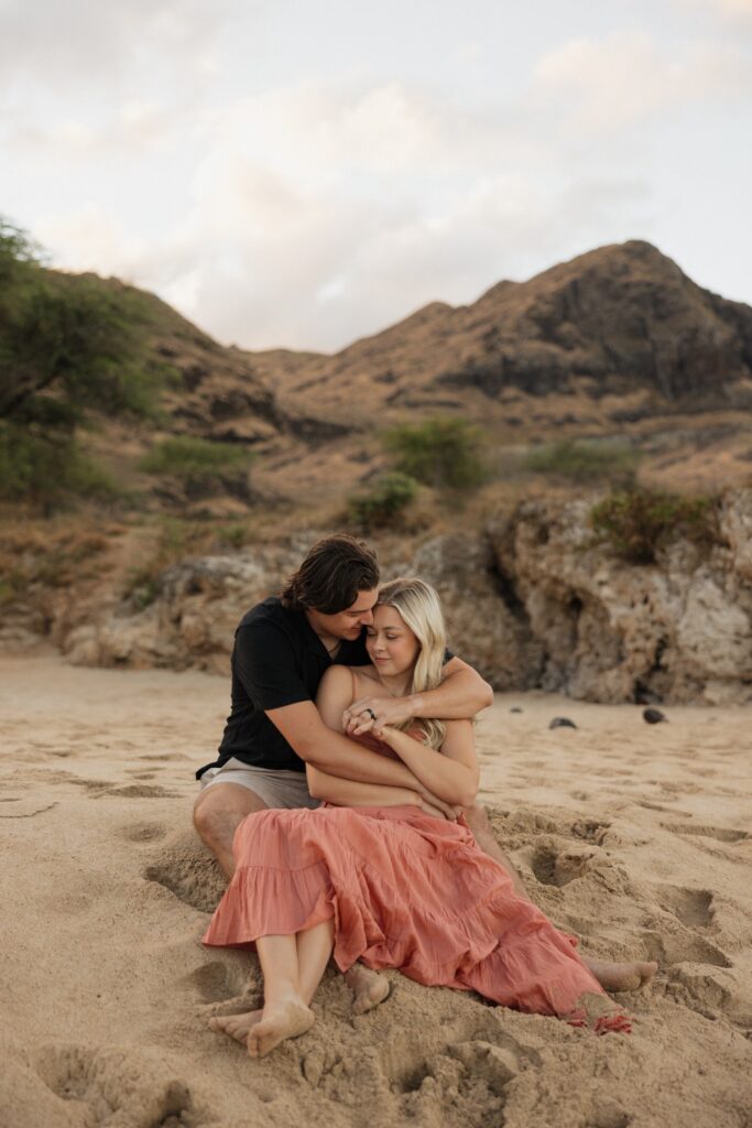 A man hugs a woman from behind while the two of them sit together on a beach with a rocky hill behind them
