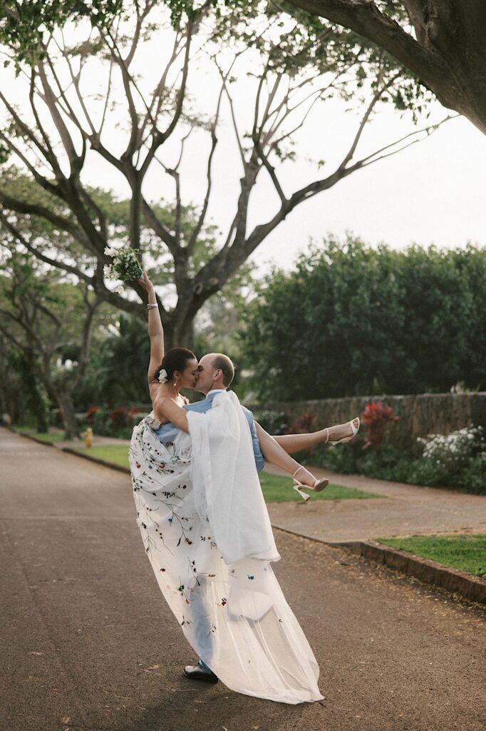 A woman in a floral wedding dress is kissed by a man as he carries her down the street and she raises her bouquet in the air