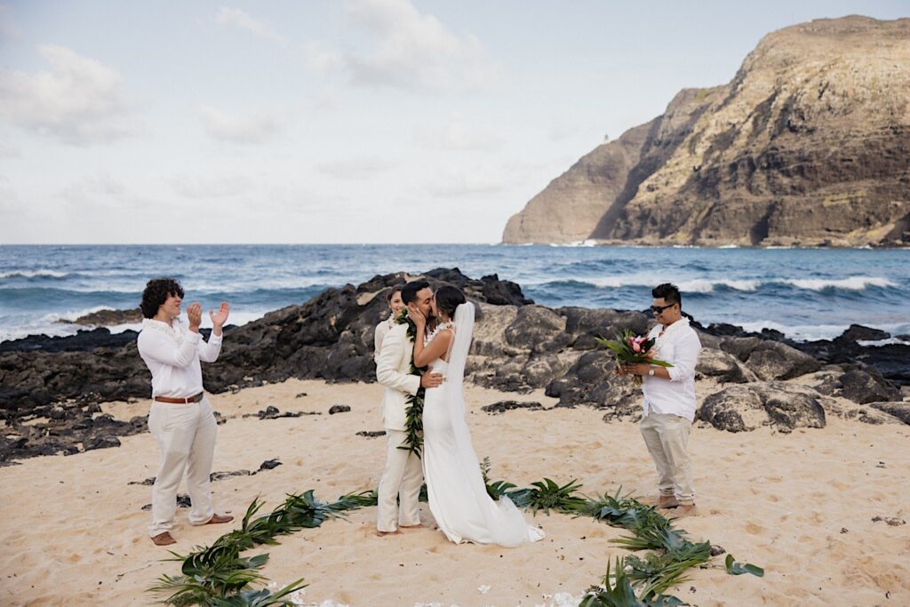 A bride and groom kiss one another during their wedding ceremony on a  beach of Oahu as guests cheer around them