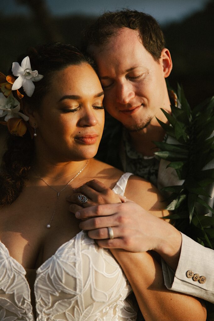 A bride is hugged from behind by the groom as the two stand with their eyes closed while the sun sets