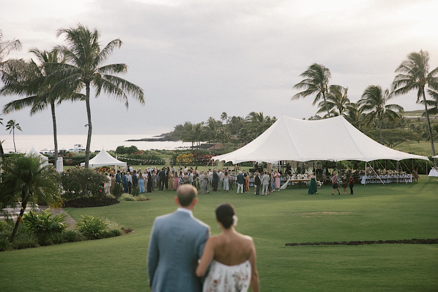A bride and groom face away from the camera and look out over a grass clearing towards the ocean and their outdoor tent wedding reception at their venue Kukui'ula