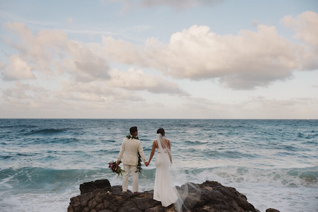 A bride and groom hold hands as they stand on a rock formation looking out at the ocean