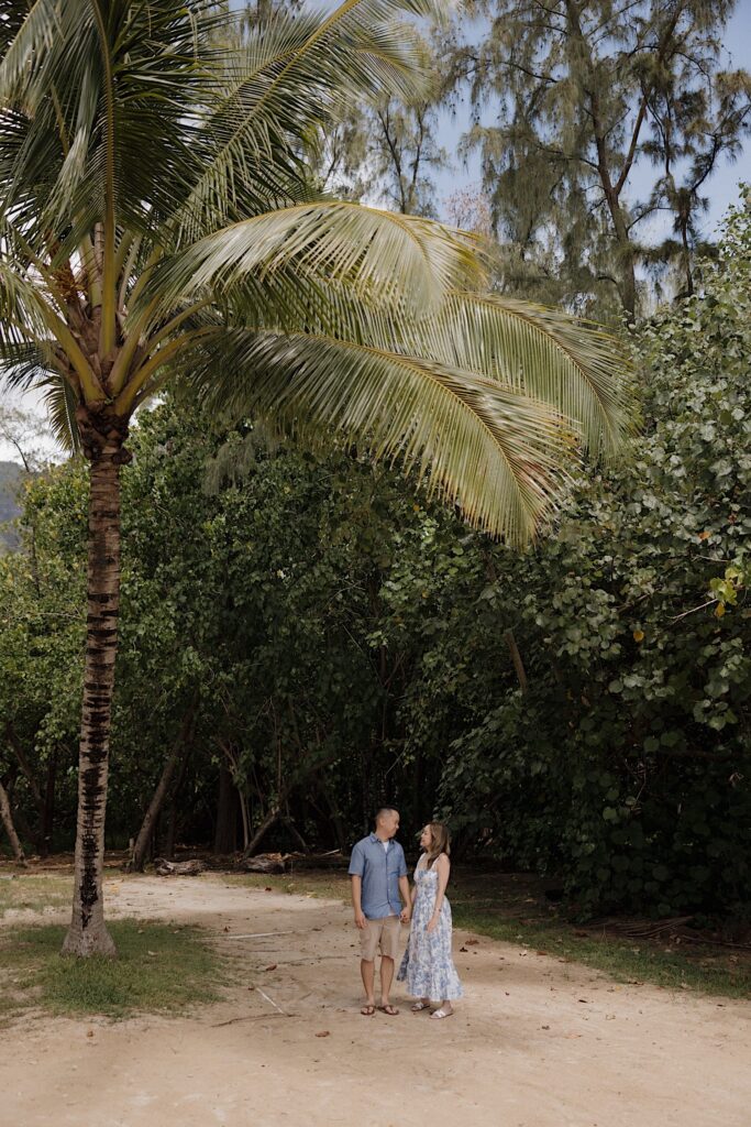 A man and woman stand holding hands underneath a palm tree