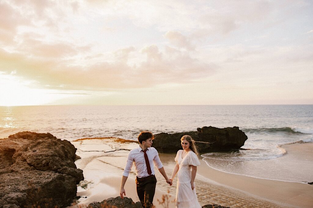 A bride is led by the hand by the groom towards the camera away from the ocean as the sun sets behind them