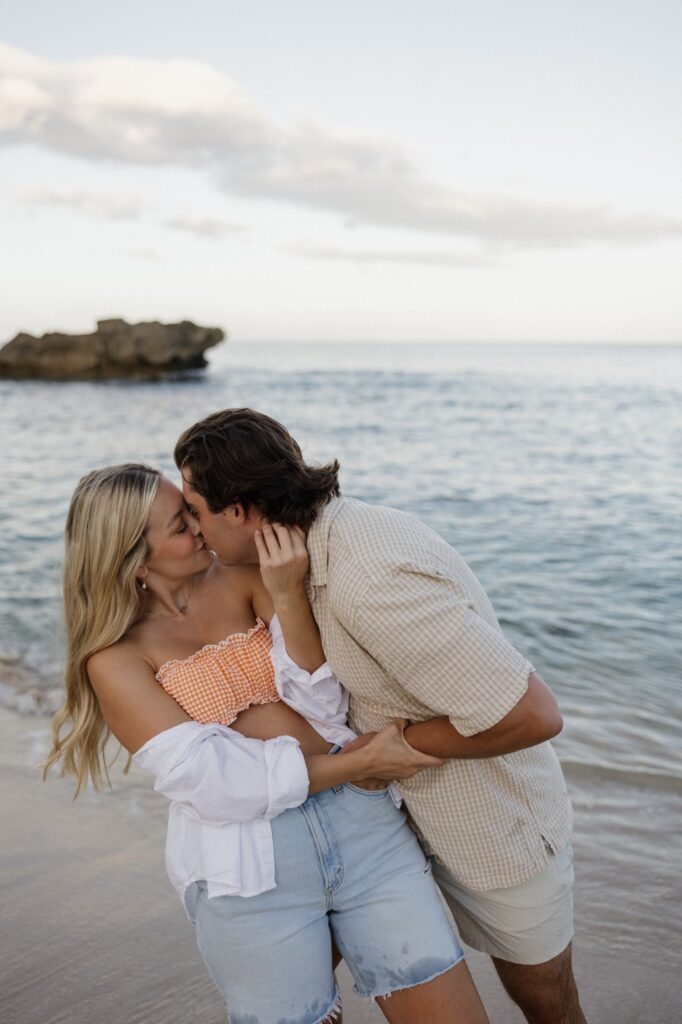 A man and woman kiss one another while standing on a beach together with the ocean behind them and a rock formation out in the water