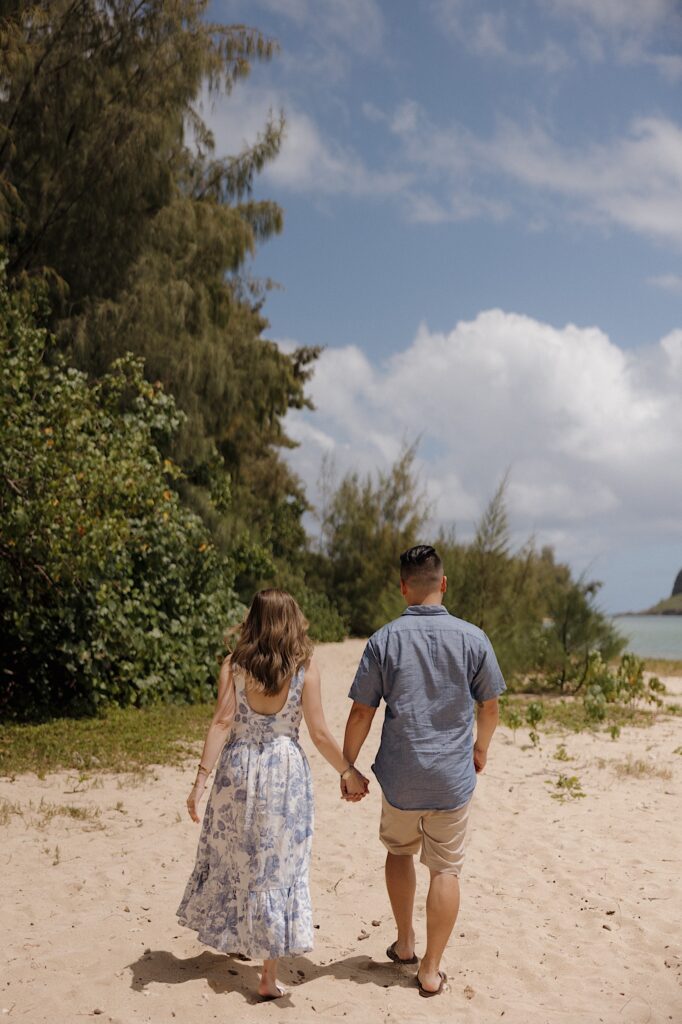 A man and woman walk hand in hand on a beach away from the camera