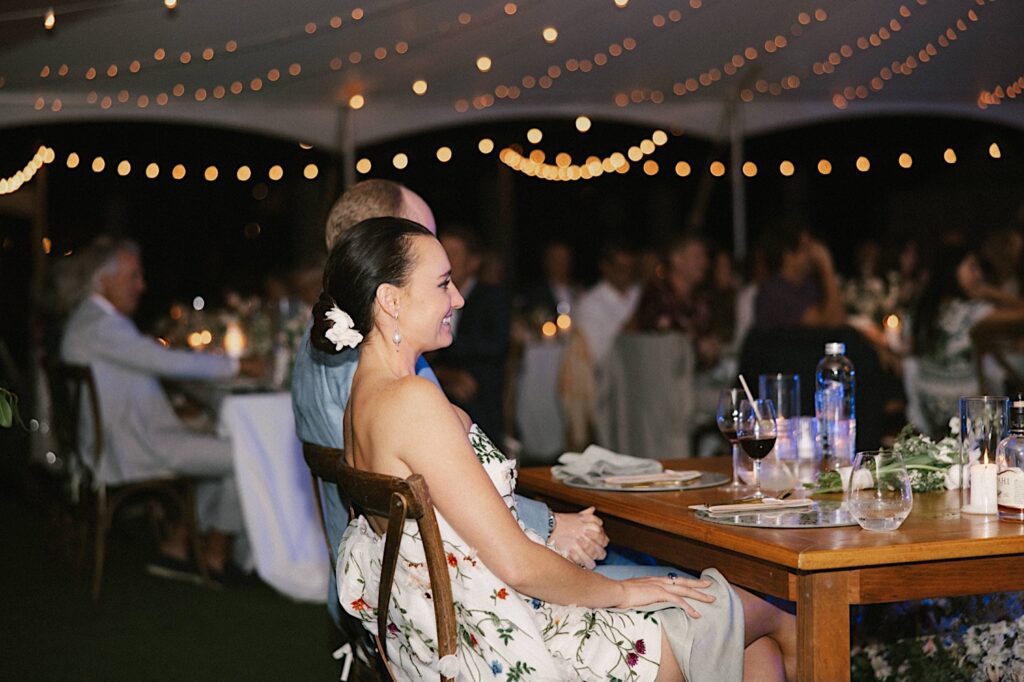 A bride and groom sit side by side at their table underneath a string lit tent during their wedding reception at their venue Kukui'ula