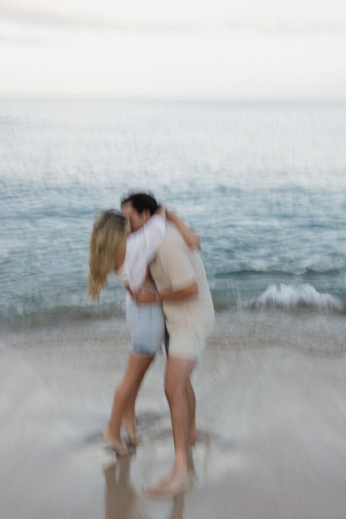 Blurry photo of a man and woman kissing one another while on a beach with the water of the ocean behind them