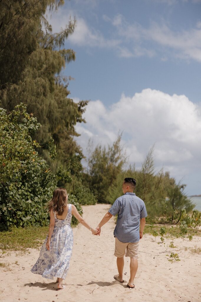 A man and woman walk hand in hand on a beach away from the camera
