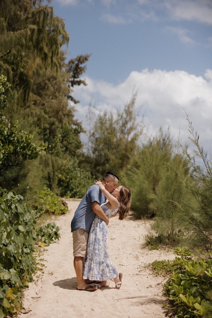 A man and woman kiss one another while standing on a sandy path surrounded by trees and shrubs