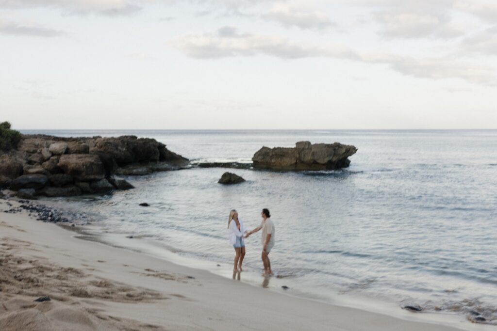 Blurry photo of a man and woman holding hands while on a beach of Oahu at sunrise during their engagement session, behind them is the ocean and rock formations in the water