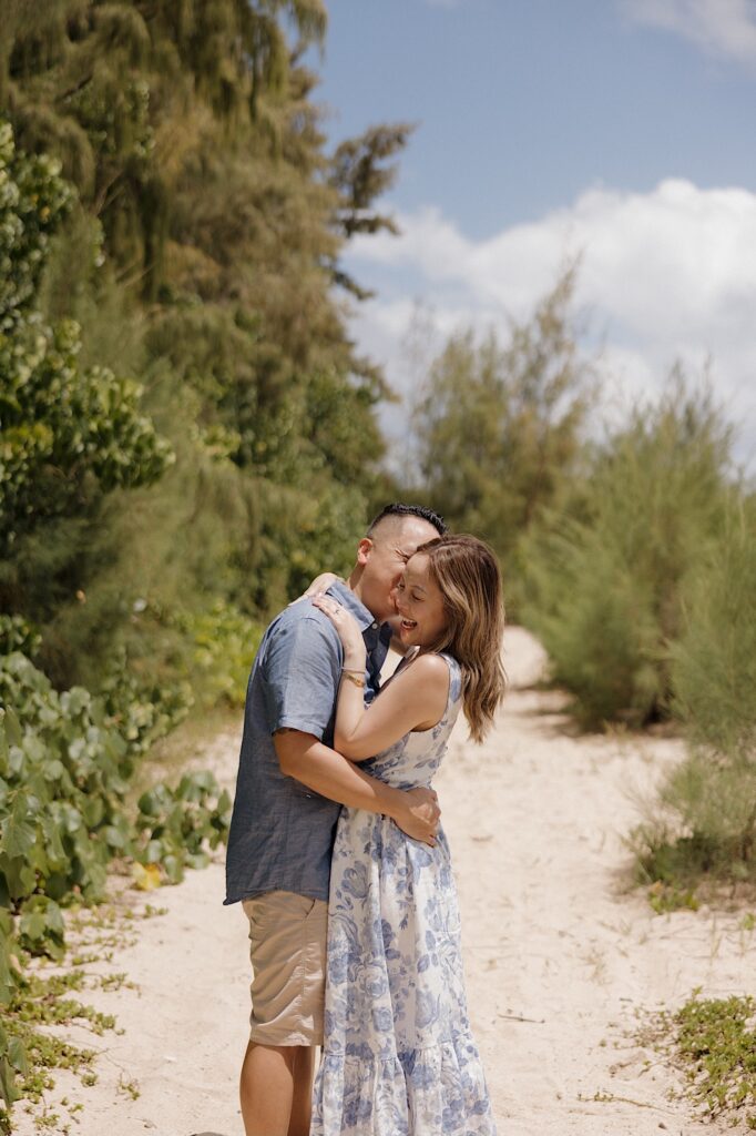 A woman smiles while a man hugs and kisses her as they stand on a sandy path