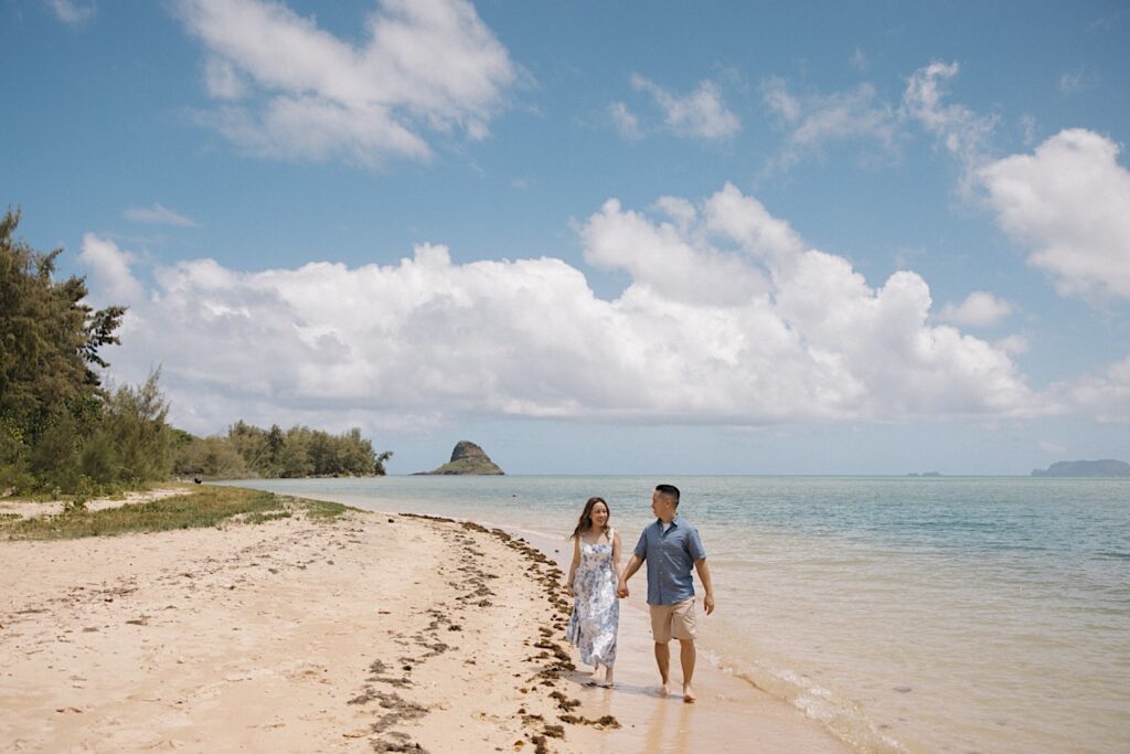 A man and woman walk along a beach on Oahu after their surprise proposal, they're holding hands and smiling at one another