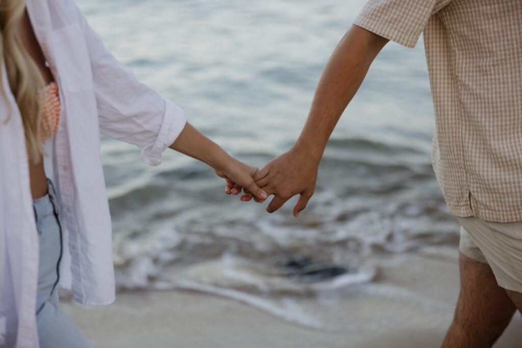 A man and woman hold hands while walking along a beach of Oahu during their sunrise engagement session