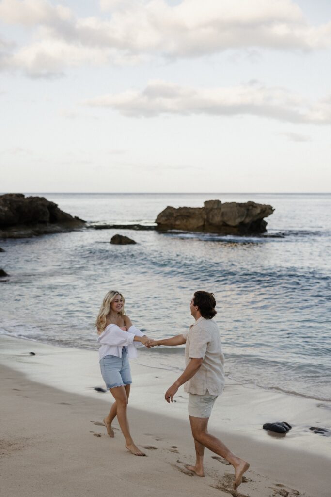 A woman holds a mans hand and drags him towards her while the two walk along a beach together with the ocean and rock formations in the water behind them
