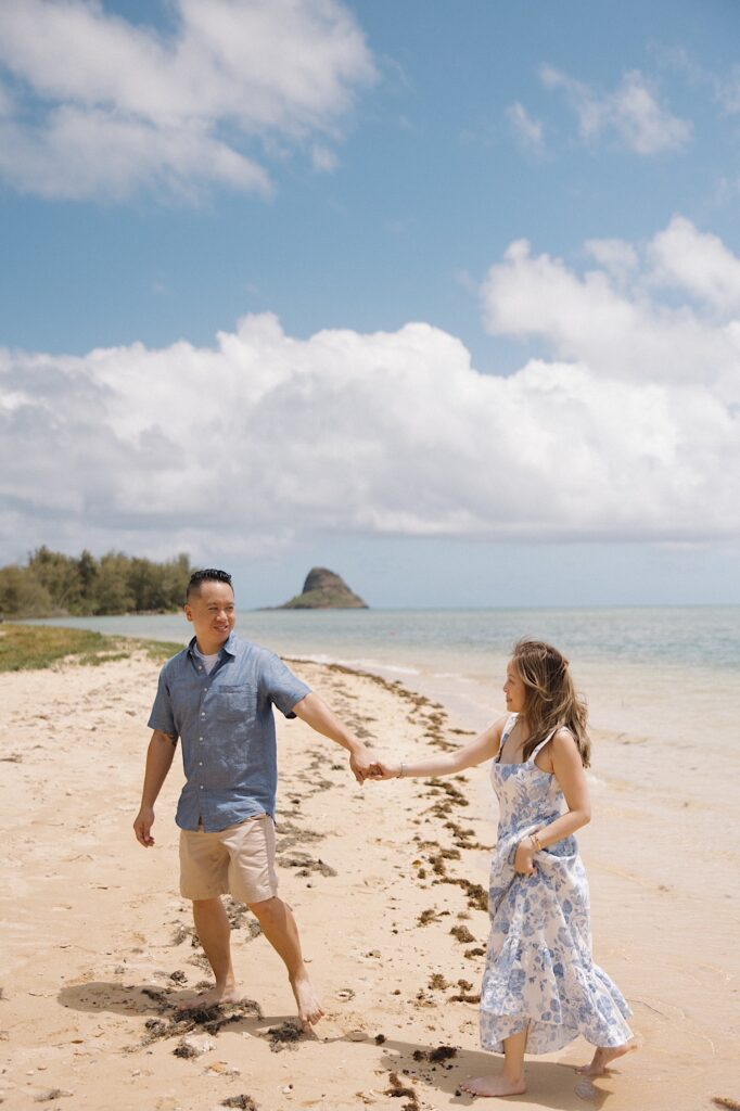 A man and woman hold hands as they walk along a beach on Oahu and smile at one another