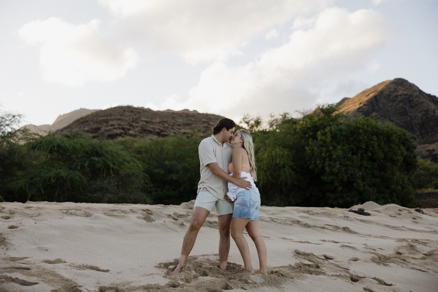 A man and woman stand on a beach on Oahu with mountains behind them, they're about to kiss one another during their engagement session taking place at sunrise