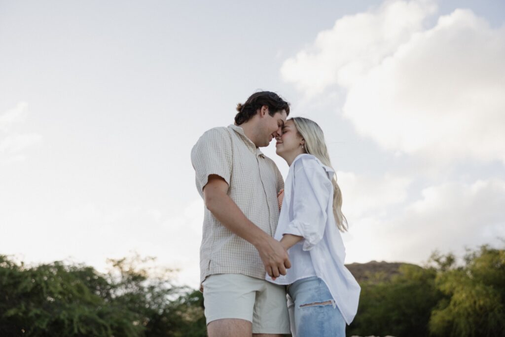 During their sunrise engagement session on a beach of Oahu a man and woman hold hands and are about to kiss one another with trees standing behind them