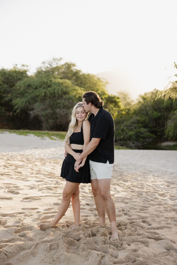 A man hugs a woman from behind and kisses her on the head while they hold hands standing together on a beach