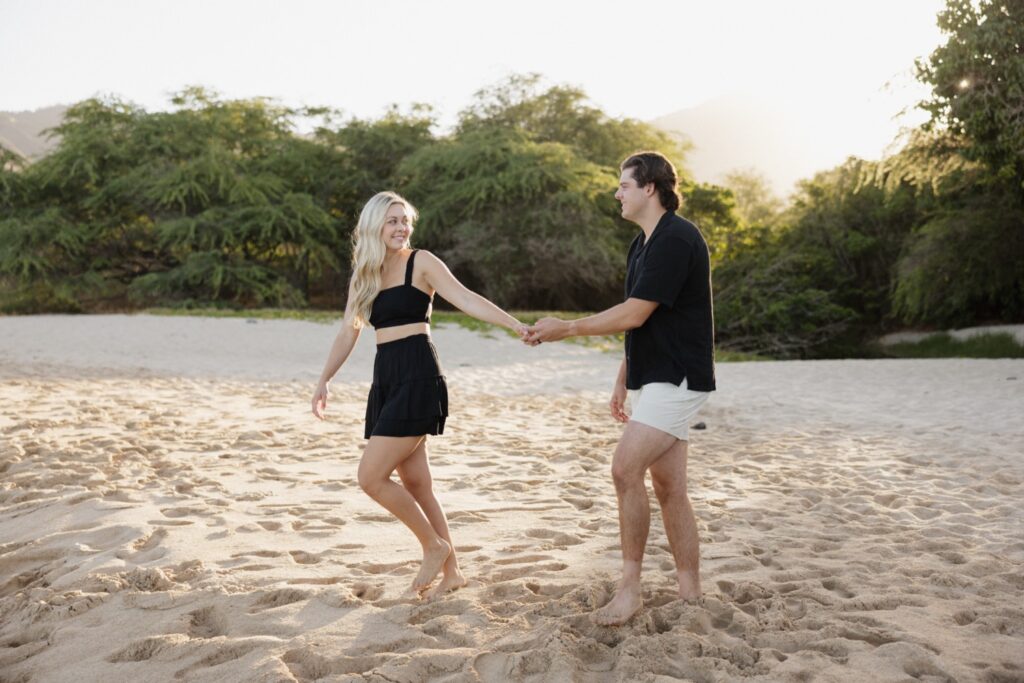 During their sunrise engagement session on a beach of Oahu a woman smiles back as she holds the hand of a man and leads him towards her