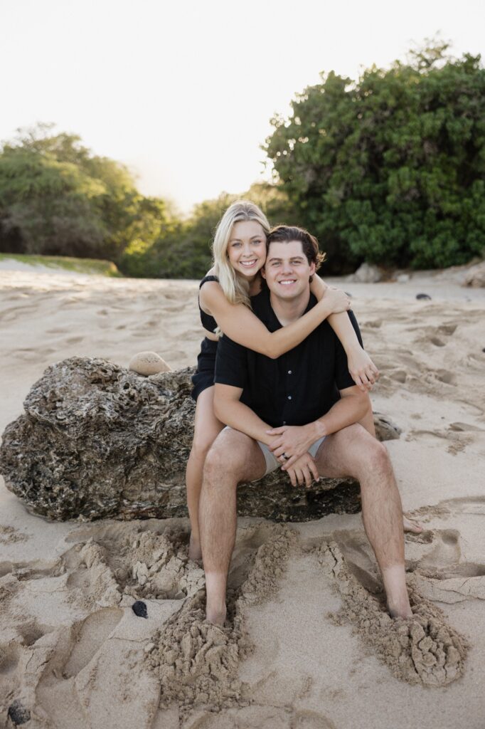 A man and woman smile at the camera while sitting together on a rock on the beach with the woman sitting behind the man and wrapping her arms around him