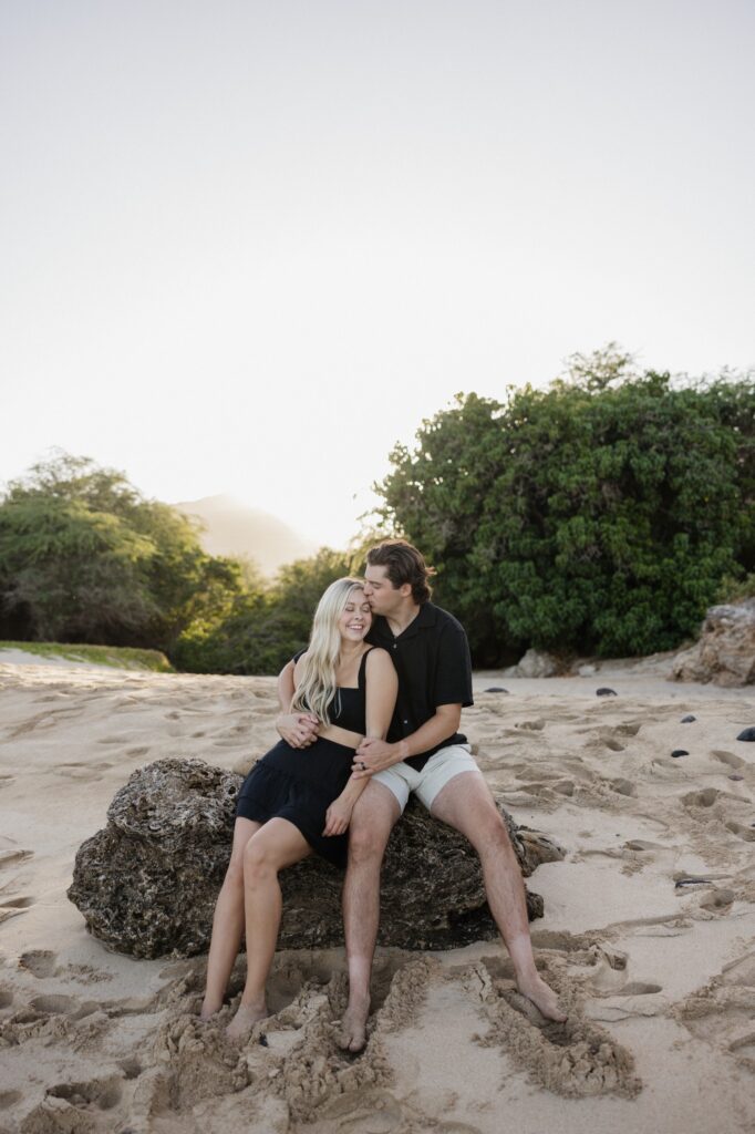 A man and woman sit together on a rock on a beach, the man hugs and kisses the woman on her head as she smiles