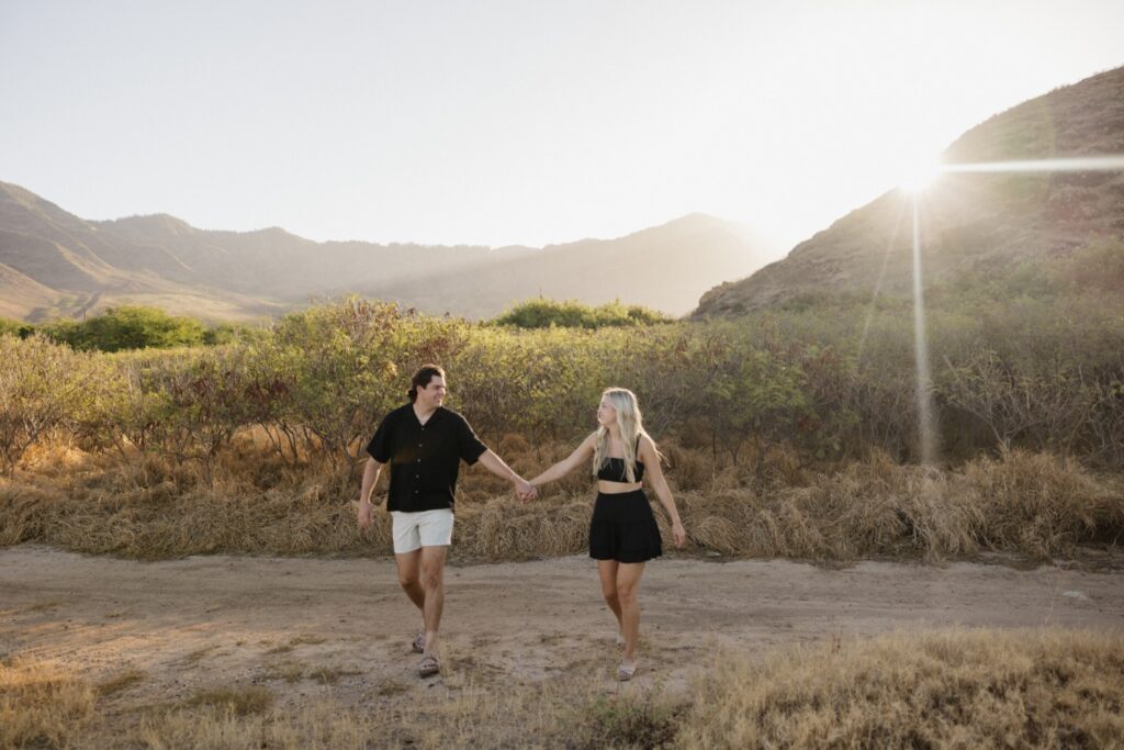 During their sunrise engagement session on Oahu a man and woman hold hands and smile at one another while walking towards the beach and the camera as the sun rises over the mountains behind them