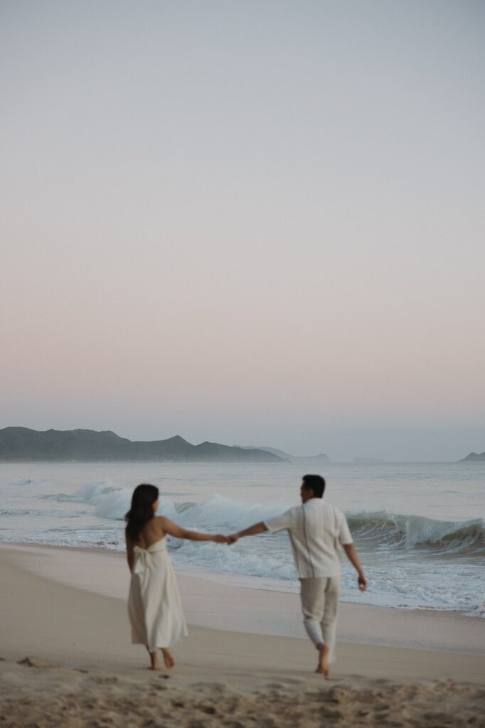 A couple walk hand in hand along Waimanalo Beach on Oahu at sunrise while waves crash along the shore