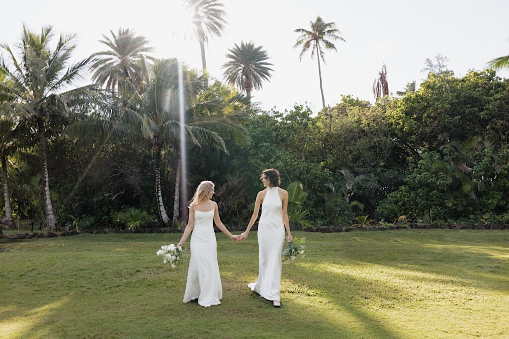 Two brides walk hand in hand towards the camera on the grounds of their wedding venue Loulu Palm on Oahu prior to their LGBTQ wedding