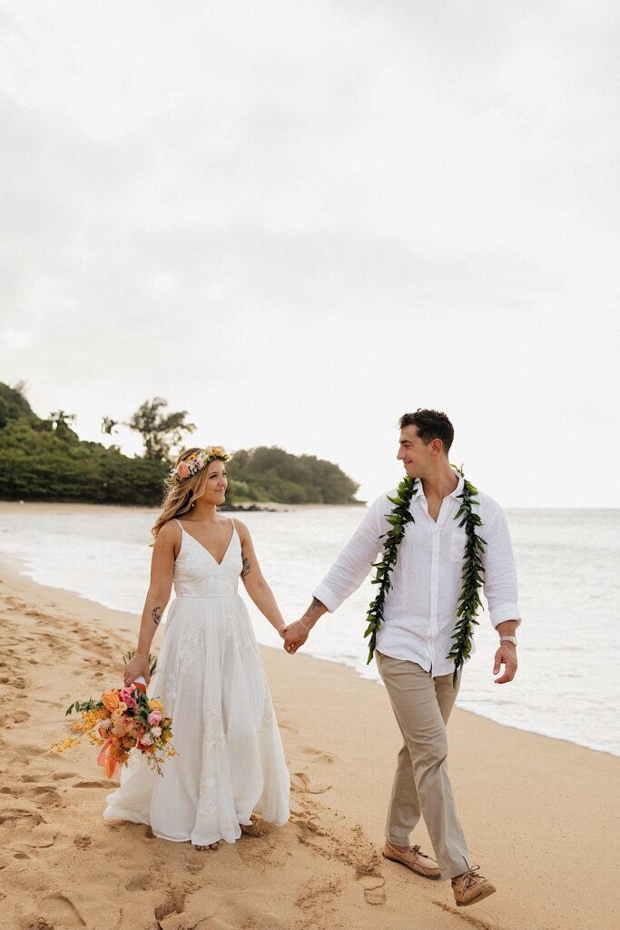 A bride and groom walk along a beach in Hawaii together as they smile at each other 